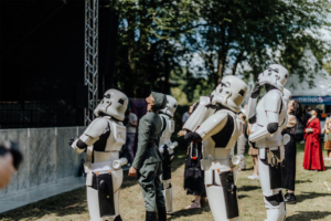 K-Pop-Dance Eine Gruppe von Stormtroopern und einem Offizier schaut gebannt zur Bühne beim Elbenwald Festival. Die Teilnehmer sind in detailreichen Kostümen gekleidet, umgeben von einer lebhaften Festivalatmosphäre mit Bäumen und Zuschauern im Hintergrund.