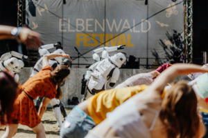 K-Pop-Dance Zwei Stormtrooper tanzen auf dem Elbenwald Festival, während Festivalbesucher in bunten Outfits mit ihnen interagieren. Im Hintergrund ist die Festivalbühne mit dem Logo gut sichtbar. Die Szene vermittelt eine lebendige, fröhliche Atmosphäre.