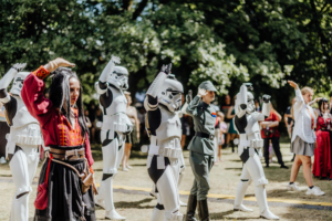 K-Pop-Dance Cosplay-Teilnehmer beim Elbenwald Festival zeigen eine beeindruckende Choreografie. Im Vordergrund eine Person in traditioneller Kleidung, umgeben von Stormtrooper-Kostümen und einem Militärcharakter, während Zuschauer im Hintergrund die Darbietung beobachten.