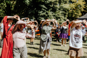K-Pop-Dance Teilnehmer des Elbenwald Festivals tanzen und feiern im Freien. Die bunte Mischung aus Kostümen und fröhlicher Stimmung spiegelt die kreative Atmosphäre des Festivals wider, während sie gemeinsam eine bewegte Performance genießen.