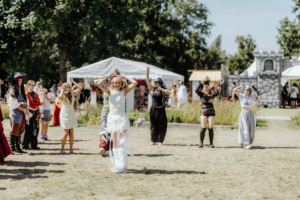 K-Pop Random Dance Tanzende Festivalbesucher in kreativen Kostümen beim Elbenwald Festival, umgeben von einer lebhaften Atmosphäre und bunten Ständen. Die Teilnehmer zeigen ihre Freude und Begeisterung in einer sommerlichen Freiluftumgebung.