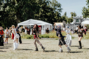 K-Pop Random Dance Tanzende Cosplayer in bunten Kostümen beim Elbenwald Festival, umgeben von Besuchern und einem Verkaufsstand. Die fröhliche Atmosphäre und kreative Kostüme spiegeln die Vielfalt der nerdigen Kultur wider.