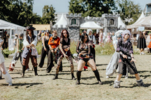 K-Pop Random Dance Gruppen von Cosplayern tanzen auf dem Elbenwald Festival, gekleidet in kreative und fantasievolle Kostüme, während im Hintergrund Festivalzelte und eine mittelalterliche Kulisse zu sehen sind. Die Atmosphäre ist lebhaft und feierlich.