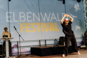 K-Pop-Dance Ein Tänzer in sportlicher Kleidung und einer roten Kappe performt auf der Bühne des Elbenwald Festivals. Im Hintergrund ist das Festival-Logo sichtbar, das eine lebhafte Atmosphäre für das nerdige Musik- und Kultur-Event schafft.