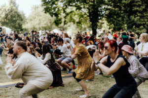K-Pop-Dance Teilnehmer des Elbenwald Festivals führen eine Gruppenübung im Freien durch, umgeben von einer begeisterten Menge. Die Veranstaltung kombiniert Musik und Nerd-Kultur, während die Atmosphäre lebhaft und einladend ist.