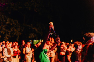 Ein begeisterter Festivalbesucher hebt sein Trinkhorn während einer Live-Performance auf dem Elbenwald Festival. Im Hintergrund jubelnde Menge, umgeben von bunten Lichtern und einer festlichen Atmosphäre, die Musik und Gemeinschaft feiert.