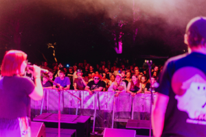 Karaoke Bühne beim Elbenwald Festival mit einer Sängerin, die vor einem begeisterten Publikum auftritt. Die Zuschauer, in nerdigen Outfits, zeigen ihre Begeisterung für die Musik und Atmosphäre des Festivals, umgeben von bunten Lichtern und einer lebhaften Stimmung.