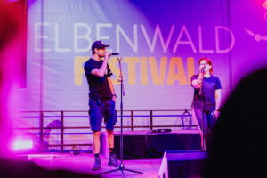 Karaoke Ein männlicher und ein weiblicher Moderator stehen auf der Bühne des Elbenwald Festivals, während sie Mikrofone halten. Im Hintergrund ist das Festival-Logo gut sichtbar. Die Beleuchtung in lila und pink schafft eine lebendige Atmosphäre.