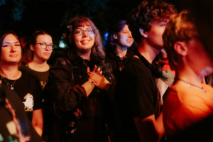 Eine Gruppe von Festivalbesuchern beim Elbenwald Festival, die begeistert auf die Bühne schauen. Die Atmosphäre ist lebhaft und die Teilnehmer tragen nerdige T-Shirts, während sie sich auf die Musik und das Event freuen.