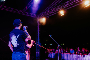 Ein Mann mit einer Crew-T-Shirt steht auf der Bühne und spricht ins Mikrofon, während eine Sängerin im Hintergrund performt. Die beleuchtete Bühne zieht eine begeisterte Menge an, die gespannt zuschaut. Elbenwald Festival, Musik und Nerdkultur vereint.