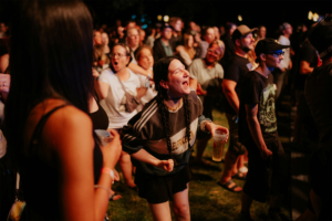 Menschenmenge beim Elbenwald Festival, begeisterte Fans singen und tanzen zur Musik. Eine junge Frau mit Zopf und sportlichem Outfit hebt ihre Hände und genießt die Atmosphäre. Im Hintergrund sind weitere Festivalbesucher zu sehen, die ebenfalls begeistert mitfeiern.