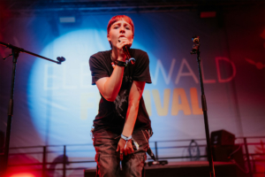Junge Musikerin auf der Bühne des Elbenwald Festivals, umgeben von bunten Lichtern. Sie singt leidenschaftlich ins Mikrofon, während das Festival-Logo im Hintergrund leuchtet. Die Atmosphäre ist lebhaft und energiegeladen, ideal für Fans von Musik und Nerd-Kultur.