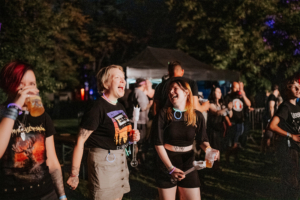 Zwei Frauen lachen und genießen Getränke beim Elbenwald Festival, umgeben von fröhlichen Festivalbesuchern. Die Szene zeigt die lebendige Atmosphäre und die bunte Mischung aus Musik und Nerd-Kultur in einer nächtlichen Umgebung.