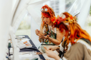 Zwei kreativ gekleidete Frauen mit roten Haaren arbeiten an einem Basteltisch beim Elbenwald Festival. Der Tisch ist mit verschiedenen Bastelmaterialien und Malutensilien dekoriert, während die Atmosphäre des Festivals im Hintergrund sichtbar ist.