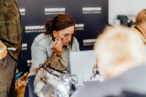 Crafting Village Eine Person mit blutiger Schminke arbeitet konzentriert an einem Makeup-Projekt am Elbenwald Festival, umgeben von anderen Künstlern und einem Tisch voller Make-up-Zubehör. Der Hintergrund zeigt das Branding des Festivals, das eine kreative Atmosphäre fördert.