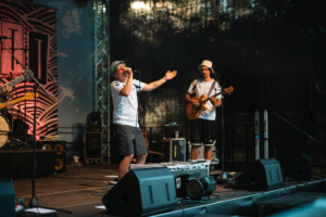 Liedfett Live-Auftritt auf dem Elbenwald Festival: Ein Sänger mit Sonnenbrille und Hut begeistert das Publikum, während ein Gitarrist im Hintergrund spielt. Die Bühne ist mit einem bunten Banner dekoriert, das die kreative Atmosphäre des Festivals unterstreicht.