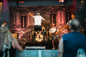 Liedfett Musiker auf der Bühne beim Elbenwald Festival, das eine Mischung aus Musikfestival und Nerd-Convention bietet. Der Frontmann springt energetisch, während die Bandmitglieder mit Instrumenten im Hintergrund spielen. Festivalbesucher genießen die Atmosphäre.
