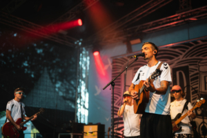 Liedfett Ein Musiker mit Gitarre steht im Mittelpunkt einer Bühnenaufführung beim Elbenwald Festival, umgeben von Bandkollegen. Die Bühne ist mit bunten Lichtern beleuchtet, während das Publikum die mitreißende Atmosphäre eines einzigartigen Musik- und Nerd-Events genießt.