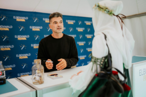 Akram El-Bahay Ein Mann spricht am Elbenwald Festival mit einer Besucher*in, die ein Kostüm trägt. Im Hintergrund ist ein Banner mit dem Festival-Logo zu sehen. Auf dem Tisch stehen eine Flasche und einige Flyer. Die Atmosphäre ist freundlich und einladend.