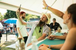 Baumann Bergmann Pokinsson Ein Besucher mit Strohhut macht ein Selfie am Stand des Elbenwald Festivals, umgeben von fröhlichen Menschen. Im Hintergrund sind Festivalbesucher und eine blaue Sonnenschirm zu sehen, die die entspannte Atmosphäre des Events unterstreichen.