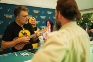 Baumann Bergmann Pokinsson Ein Musiker spielt auf einer Ukulele beim Elbenwald Festival, umgeben von verkleideten Fans. Im Hintergrund sind weitere Festivalbesucher zu sehen, die gespannt zuschauen. Bunte Stände und eine festliche Atmosphäre prägen die Szene.