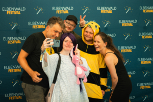 Baumann Bergmann Pokinsson Eine Gruppe von fünf Personen posiert fröhlich für ein Selfie vor dem Hintergrund des Elbenwald Festivals, gekleidet in verschiedenen Kostümen, darunter ein Bienenanzug und ein Bunny-Plüschspielzeug. Die Atmosphäre ist ausgelassen und feierlich.