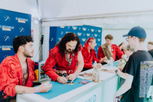 Corvus Corax Schauspieler im roten Kostüm signieren Autogramme am Elbenwald Festival, während Fans in Warteschlange stehen. Im Hintergrund sind Banner mit dem Festival-Logo sichtbar, die die nerdige Atmosphäre des Events unterstreichen.