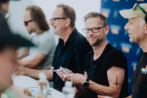 Fiddler's Green Eine Gruppe von Personen sitzt an einem Tisch auf dem Elbenwald Festival. Einer der Männer, mit Brille und Bart, lächelt in die Kamera, während er einen Stift in der Hand hält. Im Hintergrund sind weitere Teilnehmer zu sehen, die sich unterhalten.