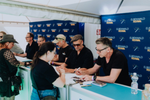 Fiddler's Green Fans stehen am Signiertisch beim Elbenwald Festival, um Autogramme von mehreren Künstlern zu erhalten. Die Atmosphäre ist entspannt und kreativ, mit einer bunten Mischung aus Nerd-Kultur und Musik.