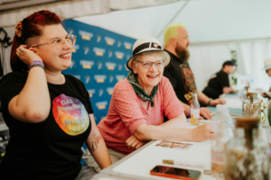 Annika Röttinger, Lisa Kuppler, Tobias M. Eckrich Zwei fröhliche Frauen mit bunten Outfits sitzen an einem Tisch, während ein Mann im Hintergrund steht. Die Szene spielt beim Elbenwald Festival, wo Fans von Musik und Popkultur zusammenkommen, um Spaß zu haben und sich auszutauschen.