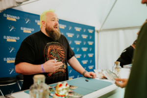 Tobias M. Eckrich Ein Teilnehmer mit leuchtend grünem Haar und Bart steht an einem Tisch beim Elbenwald Festival. Er hält einen Stift in der Hand und interagiert mit einem Fan, während im Hintergrund das Festival-Logo sichtbar ist. Merchandise-Artikel liegen auf dem Tisch.