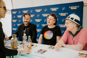 Annika Röttinger, Lisa Kuppler, Stefan Servos Ein lächelndes Trio von Gästen am Tisch beim Elbenwald Festival, umgeben von Merchandise und Getränken. Im Hintergrund ist das Festival-Logo zu sehen. Die Atmosphäre ist entspannt und freundlich, ideal für Fans von Musik und Nerdkultur.