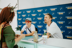 Liedfett Zwei lächelnde Mitarbeiter am Elbenwald Festival stehen hinter einem Info-Stand, während sie mit einem Besucher sprechen. Im Hintergrund ist das Festival-Logo auf einem blauen Banner sichtbar. Die Atmosphäre ist freundlich und einladend.