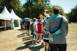 Tag der Helden Menschen stehen in einer Schlange beim Elbenwald Festival, umgeben von Zelten und Bäumen. Ein Besucher trägt ein T-Shirt mit dem Schriftzug „SUNSET STEPS“ und hält einen bunten Stock mit einer Figur in der Hand. Sonnenschein und festliche Stimmung prägen die Szene.