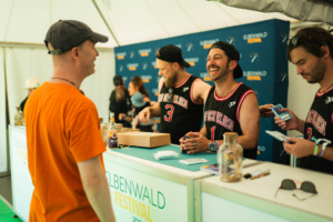 Tag der Helden Besucher interagiert am Stand des Elbenwald Festivals mit Mitarbeitern in sportlichen Trikots. Die Szene zeigt eine freundliche Atmosphäre, während Festivalbesucher Tickets und Merchandise erwerben. Hintergrund mit Festivalbranding sichtbar.