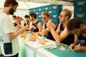 Tag der Helden Fans stehen an einem Tisch beim Elbenwald Festival, um Autogramme von Bandmitgliedern in sportlichen Trikots zu erhalten. Die Atmosphäre ist lebhaft, mit Getränken und Merchandise auf dem Tisch. Im Hintergrund sind Festivalbanner sichtbar.