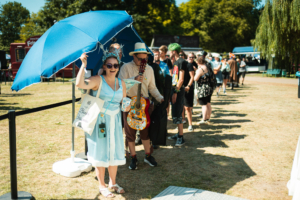 Eine fröhliche Festivalbesucherin in einem blauen Kostüm hält einen großen blauen Regenschirm und posiert lächelnd vor einer langen Warteschlange von Teilnehmern, die auf dem Elbenwald Festival stehen. Die Szene spielt sich an einem sonnigen Tag im Freien ab.