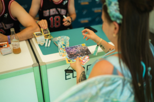 Tag der Helden Frau mit buntem Haar und Tattoos hält ein signiertes Foto in der Hand, während sie an einem Tisch mit Künstlern beim Elbenwald Festival sitzt. Auf dem Tisch sind Merchandise-Artikel und Stifte sichtbar. Die Atmosphäre ist lebhaft und kreativ.