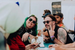 dArtagnan Zwei Teilnehmer des Elbenwald Festivals posieren lächelnd an einem Tisch. Die Frau mit grünen Haaren trägt eine Blumenkrone und Sonnenbrille, während der Mann mit tätowiertem Arm einen freundlichen Gruß zeigt. Im Hintergrund sind weitere Festivalbesucher zu sehen.