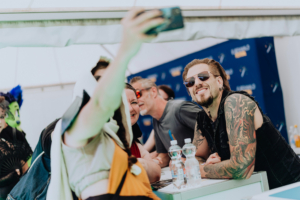 dArtagnan Selfie-Moment auf dem Elbenwald Festival: Besucher posieren mit einem Künstler, umgeben von fröhlichen Fans in kreativen Outfits. Im Hintergrund sind Festivalbanner sichtbar, die die lebendige Atmosphäre des Events unterstreichen.