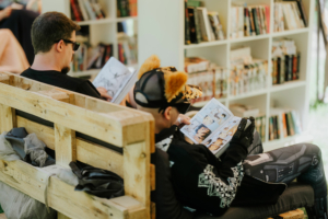 Carlsen Manga Lounge Zwei Besucher des Elbenwald Festivals entspannen auf einer Holzbank und lesen Comics. Im Hintergrund sind Bücherregale mit einer Vielzahl von Comics und Romanen sichtbar, die die nerdige Atmosphäre des Festivals unterstreichen.