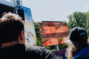 Mario Kart Turnier-Finale Zuschauer beim Elbenwald Festival beobachten ein spannendes Videospielturnier auf einer Großbildleinwand. Die Teilnehmer spielen ein beliebtes Rennspiel, während die Festivalatmosphäre im Hintergrund lebhaft ist.