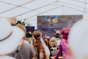 Mario Kart Turnier Besucher des Elbenwald Festivals verfolgen gebannt ein Videospielturnier auf einer großen Leinwand. Im Hintergrund ist ein DJ zu sehen, der die Stimmung anheizt, während die Teilnehmer in kreativen Kostümen zusammenkommen.