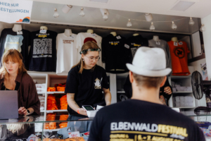 Verkaufstand beim Elbenwald Festival, mit zwei Mitarbeiterinnen, die Merchandise-Artikel wie T-Shirts und Hoodies anbieten. Die Atmosphäre ist lebhaft und einladend, perfekt für Festivalbesucher, die nach einzigartigen Souvenirs suchen.