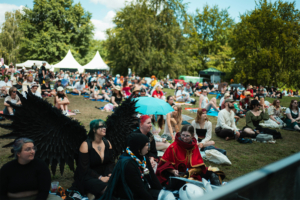 Besucher des Elbenwald Festivals entspannen auf dem Rasen, umgeben von Bäumen und Zelten. Einige tragen kreative Kostüme, darunter eine Person mit schwarzen Flügeln. Die Atmosphäre ist lebhaft und einladend, während die Menge auf die nächste Darbietung wartet.