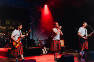 My Wife and the Microwave Drei Musiker in karierter Kleidung stehen auf der Bühne beim Elbenwald Festival. Ein Sänger mit Mikrofon und zwei weitere Musiker mit Bass und Gitarre performen vor einem bunten Lichtspiel, umgeben von Musikinstrumenten und einem lebhaften Publikum.