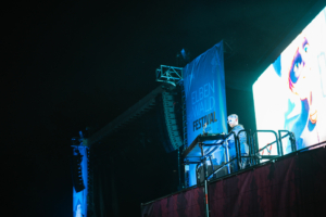 Nerds At Raves DJ auf der Bühne beim Elbenwald Festival, umgeben von beeindruckenden Lautsprechern und einem großen Bildschirm, der visuelle Effekte zeigt. Die Atmosphäre ist lebhaft und energiegeladen, während die Menge die Musik genießt.