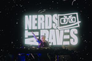 Nerds At Raves DJ auf der Bühne beim Elbenwald Festival, umgeben von einer lebhaften Atmosphäre. Im Hintergrund leuchtet das Banner "NERDS AT RAVES", das die Verbindung von Musik und Nerdkultur feiert. Farbenfrohe Lichter und ein energiegeladenes Publikum ergänzen die Szene.