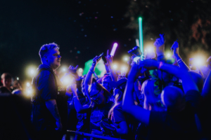 Nerds At Raves Ein Musiker steht auf der Bühne beim Elbenwald Festival, umgeben von begeisterten Fans, die mit leuchtenden Handylichtern und Schaumstoffstäben feiern. Die Atmosphäre ist energiegeladen und farbenfroh, mit vielen Menschen, die ihre Freude ausdrücken.