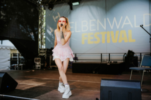 Open Stage Eine Sängerin mit kurzen blonden Haaren und roten Sonnenbrillen performt auf der Bühne des Elbenwald Festivals. Sie trägt ein rosa Outfit mit Rüschen und weiße Plattformschuhe. Im Hintergrund ist das Festival-Logo sichtbar.